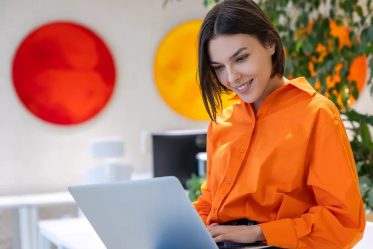 Smiling contented beautiful young dark-haired female seated on the desk typing on her portable computer